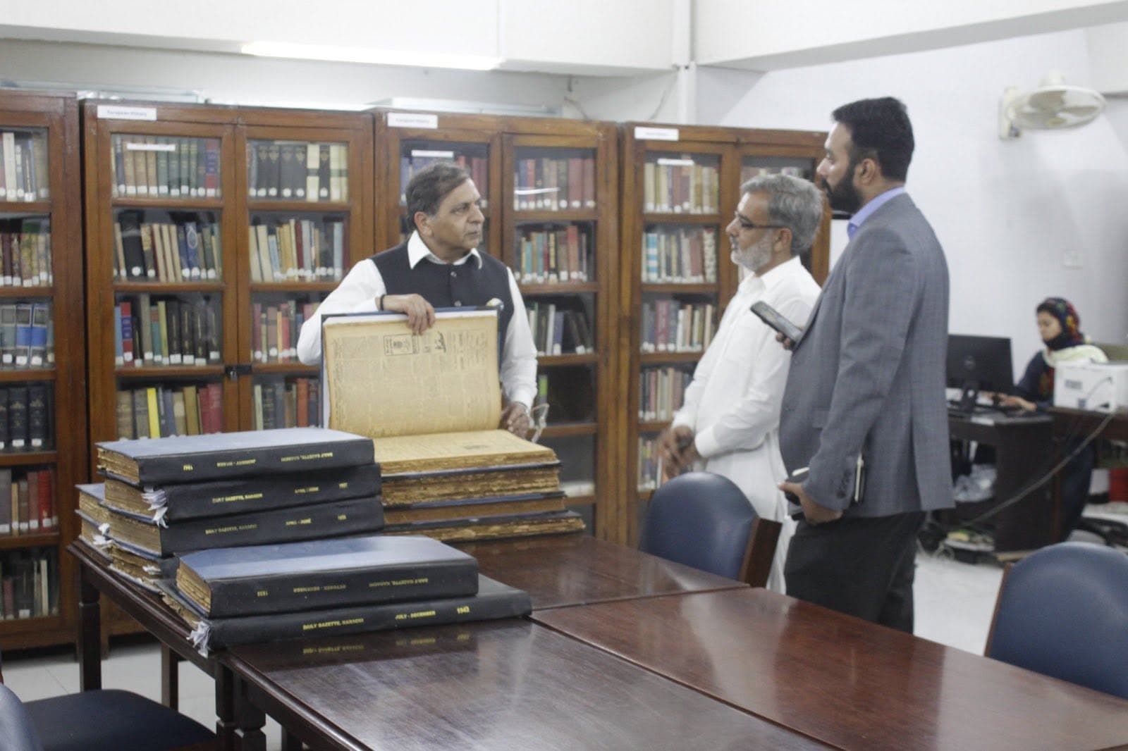 Bound volumes of the Daily Gazette, Karachi, stacked on a reading table in front of glass-fronted bookcases
