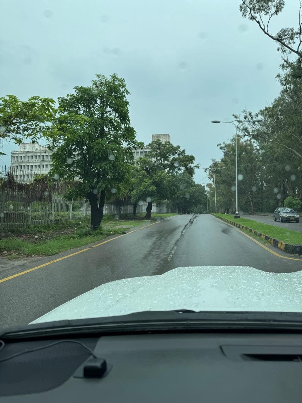 A wet, tree-lined road between white Secretariat buildings, seen through a car windscreen
