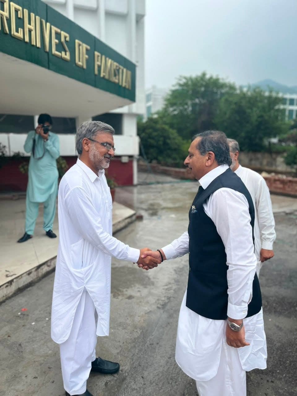 Two men shaking hands on the wet forecourt beneath the National Archives of Pakistan sign