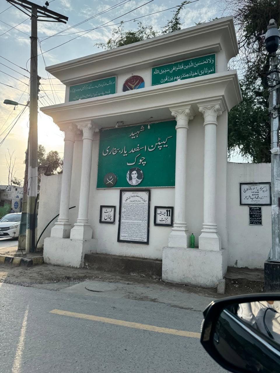 A white columned memorial with green Urdu signboards naming Shaheed Captain Asfandyar Bokhari Chowk