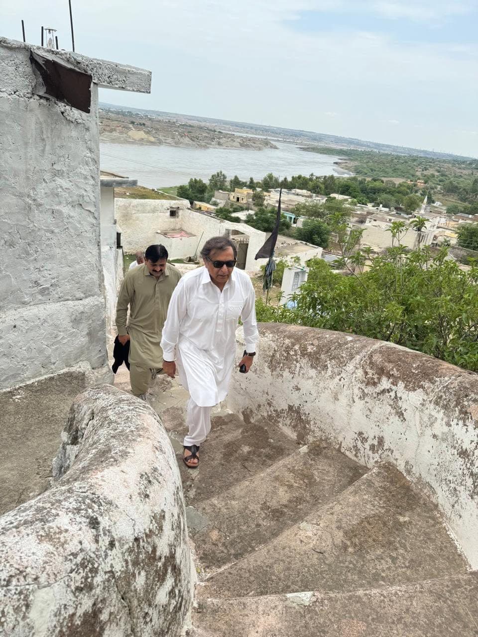 Two men climbing worn stone steps above village houses, with the wide Indus beyond