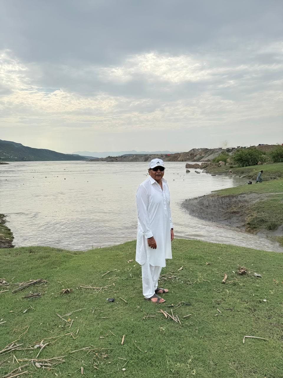 A man in white standing on a grassy bank beside the wide, silt-brown Indus under heavy cloud
