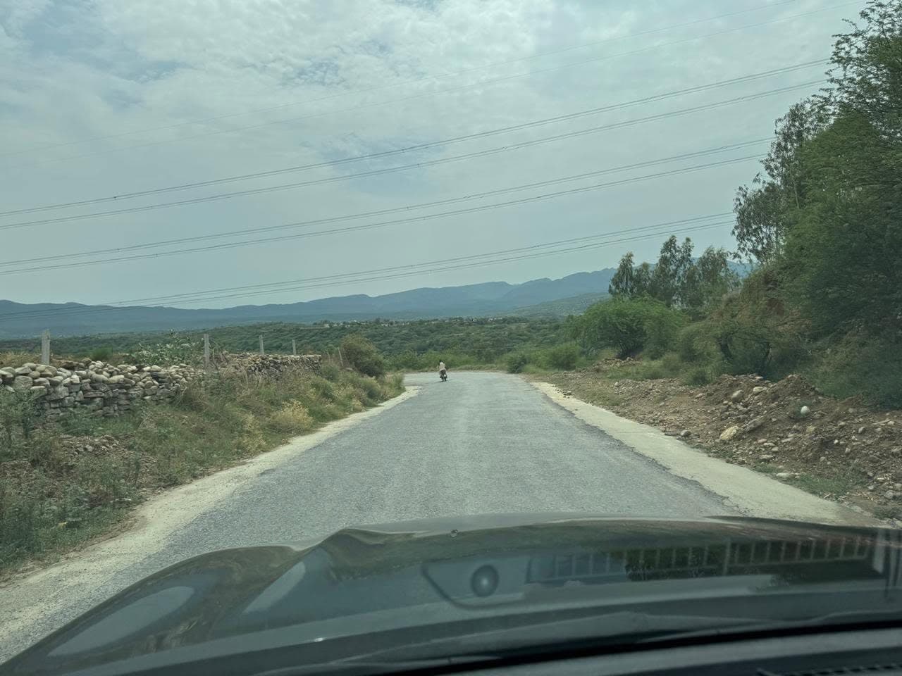 A narrow paved road running between stone walls and scrub towards distant hills, with a motorcyclist ahead