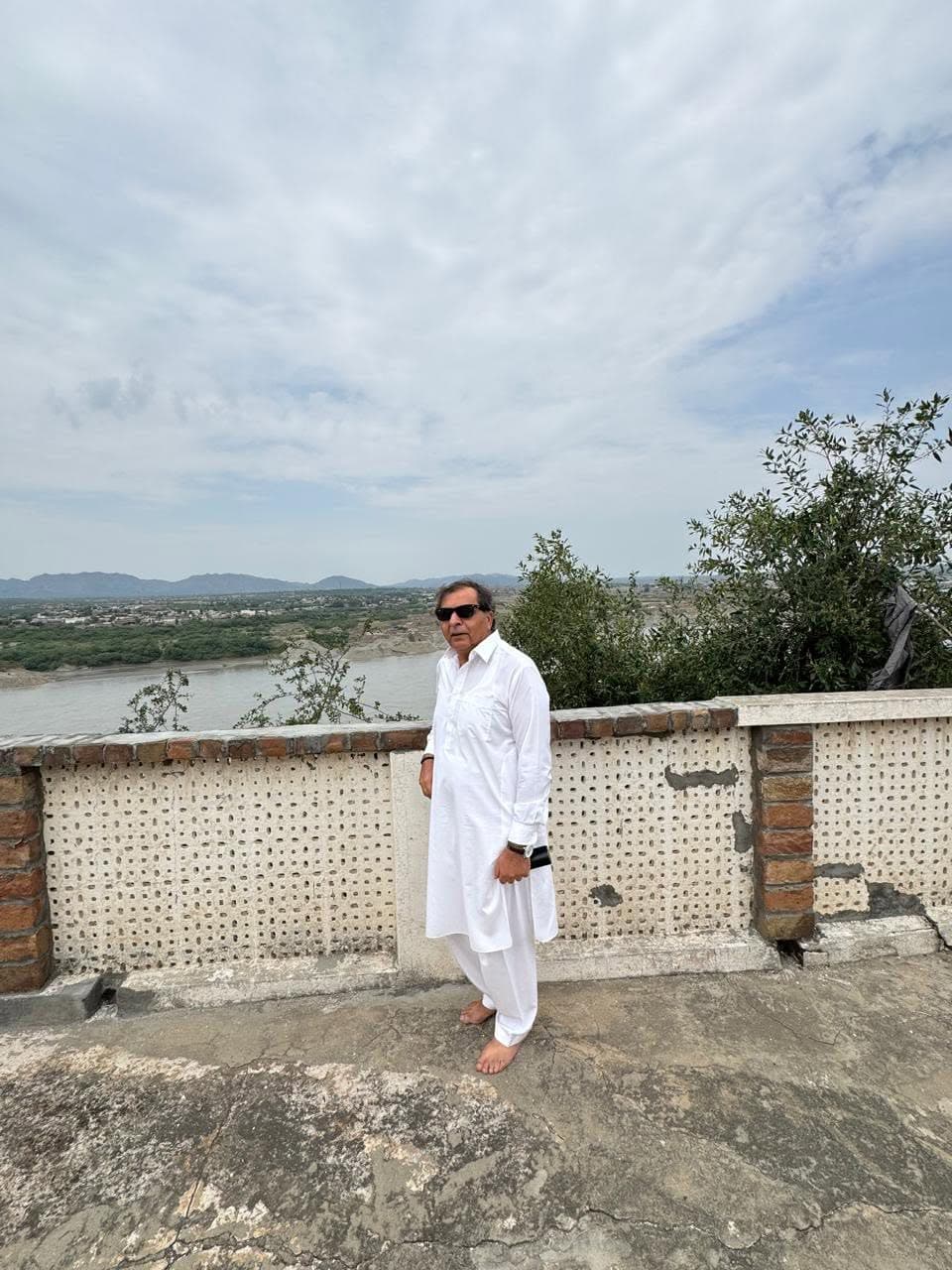 A man in white standing at a perforated parapet with the broad Indus and distant hills behind
